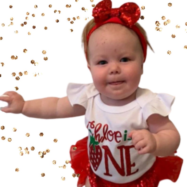 Może przedstawiać: Mała dziewczynka w białym body z czerwoną truskawką i napisem "Chloe is One" w kolorze czerwonym. Ma na sobie czerwoną opaskę na głowę z kokardką i czerwoną tiulową sp&oacute;dniczkę. Tło jest białe z złotym brokatem.