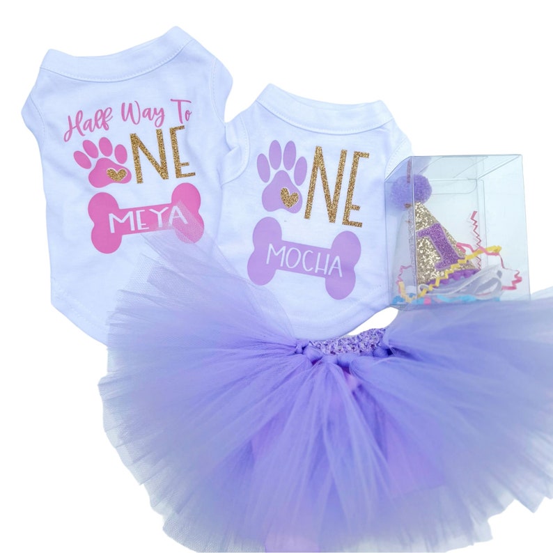 May include: Two white sleeveless pet shirts with pink and gold glitter graphics reading Half Way To ONE MEYA and ONE MOCHA, a layered lavender tulle tutu, and a gold glitter party hat with a purple number one inside a clear plastic box.