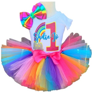 Puede incluir: Un conjunto de cumpleaños con temática de arcoíris para un niño de un año. El conjunto incluye una camiseta blanca con el nombre "Sadie Jo" y el número "1", una falda tutú colorida y un lazo a juego.