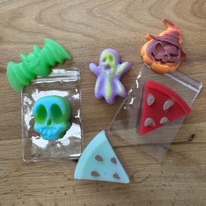 May include: A selection of Halloween-themed soaps. The soaps are shaped like a green bat, a blue and teal skull, a purple ghost, an orange pumpkin, a red pizza slice, and a blue watermelon slice. Each soap is in a clear plastic bag.
