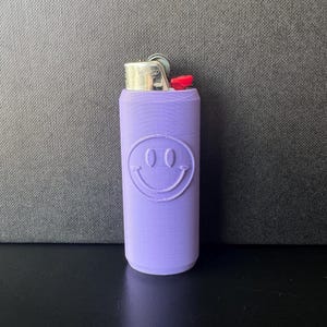 Może przedstawiać: Lawendowe etui na zapalniczkę z motywem uśmiechniętej buzi. Etui jest cylindryczne i zakrywa standardową zapalniczkę, z widoczną metalową g&oacute;rą i czerwonym przyciskiem zapłonu. Uśmiechnięta buzia ma dwa owalne oczy i zakrzywione usta.