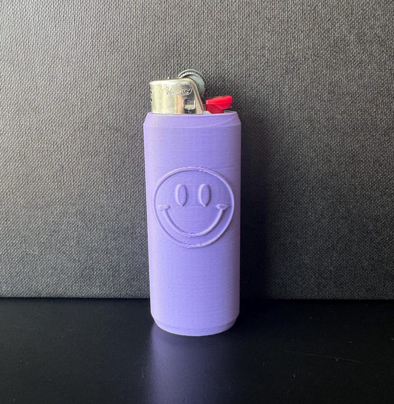 Może przedstawiać: Lawendowe etui na zapalniczkę z motywem uśmiechniętej buzi. Etui jest cylindryczne i zakrywa standardową zapalniczkę, z widoczną metalową g&oacute;rą i czerwonym przyciskiem zapłonu. Uśmiechnięta buzia ma dwa owalne oczy i zakrzywione usta.