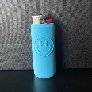 Może przedstawiać: Jasnoniebieskie etui na zapalniczkę z motywem uśmiechniętej buzi. Etui jest cylindryczne i ma srebrną metalową g&oacute;rę z czerwonym przyciskiem. Uśmiechnięta buzia jest wytłoczona z przodu.