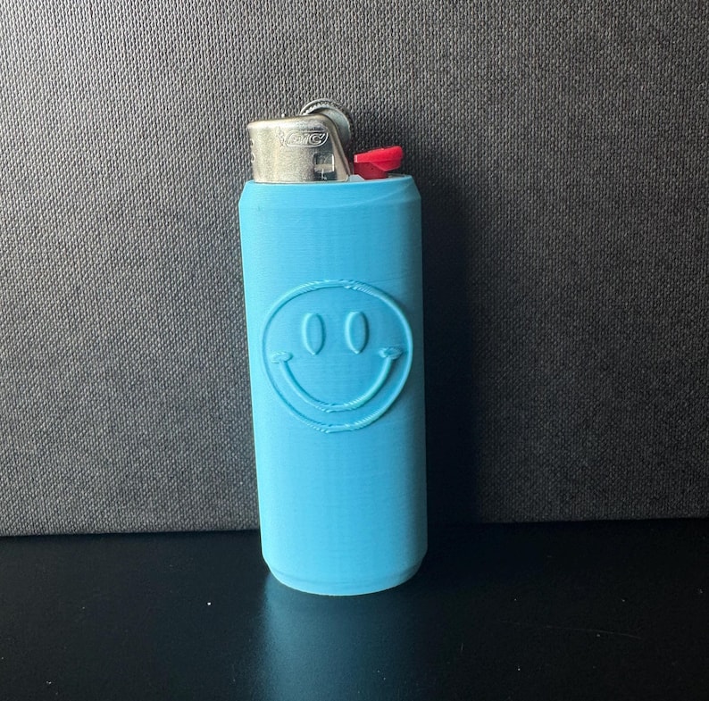 Może przedstawiać: Jasnoniebieskie etui na zapalniczkę z motywem uśmiechniętej buzi. Etui jest cylindryczne i ma srebrną metalową g&oacute;rę z czerwonym przyciskiem. Uśmiechnięta buzia jest wytłoczona z przodu.