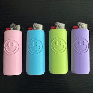 Może przedstawiać: Cztery kolorowe zapalniczki w kolorze r&oacute;żowym, niebieskim, zielonym i fioletowym. Każda zapalniczka ma wz&oacute;r uśmiechniętej buzi. Zapalniczki ułożone są w rzędzie na ciemnej powierzchni.