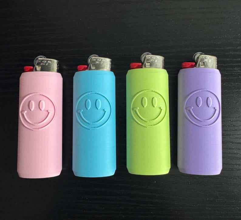 Może przedstawiać: Cztery kolorowe zapalniczki w kolorze r&oacute;żowym, niebieskim, zielonym i fioletowym. Każda zapalniczka ma wz&oacute;r uśmiechniętej buzi. Zapalniczki ułożone są w rzędzie na ciemnej powierzchni.