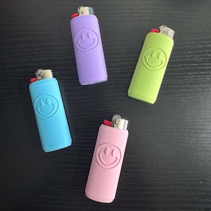 Może przedstawiać: Cztery kolorowe zapalniczki w odcieniach niebieskiego, fioletowego, zielonego i r&oacute;żowego, każda z uśmiechniętym wzorem twarzy. Zapalniczki są ułożone na ciemnej powierzchni, prezentując swoje żywe kolory i zabawny design.