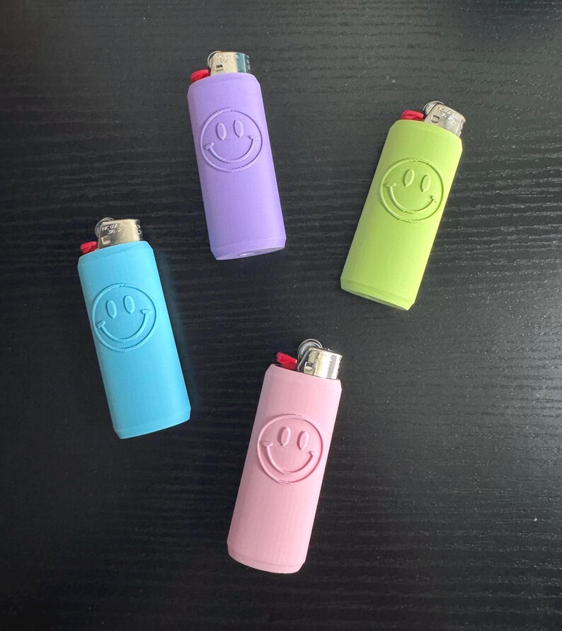 Może przedstawiać: Cztery kolorowe zapalniczki w odcieniach niebieskiego, fioletowego, zielonego i r&oacute;żowego, każda z uśmiechniętym wzorem twarzy. Zapalniczki są ułożone na ciemnej powierzchni, prezentując swoje żywe kolory i zabawny design.