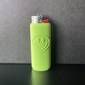 Może przedstawiać: Neonowo-zielona zapalniczka z motywem uśmiechniętej buzi. Zapalniczka ma srebrną metalową g&oacute;rę i czerwony przycisk zapłonu. Uśmiechnięta buzia jest wytłoczona z przodu.