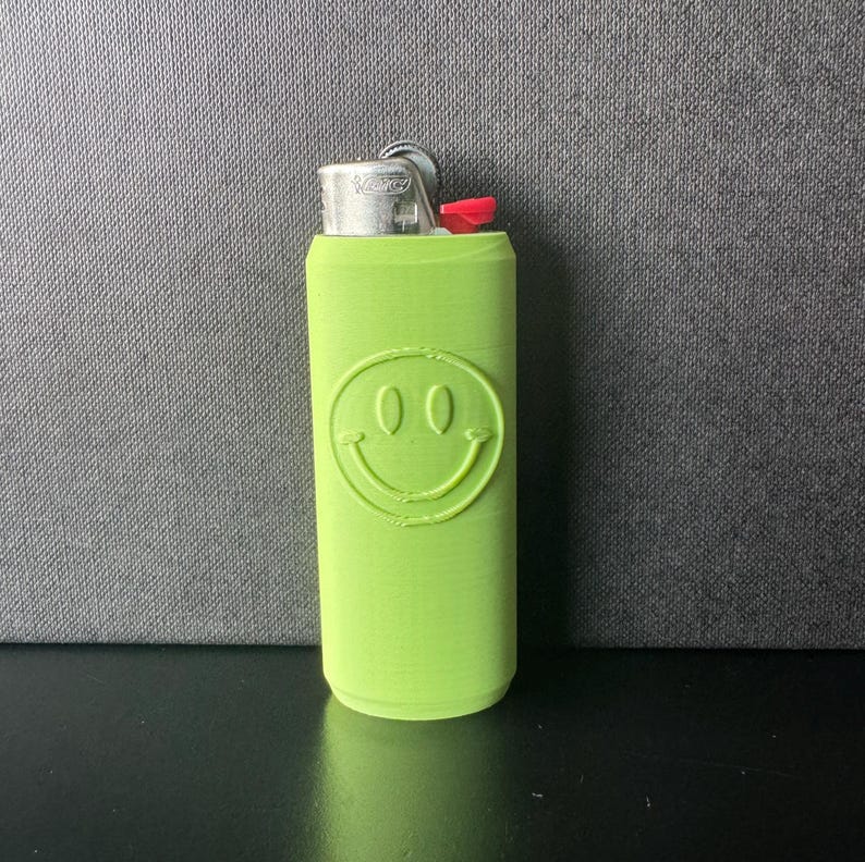 Może przedstawiać: Neonowo-zielona zapalniczka z motywem uśmiechniętej buzi. Zapalniczka ma srebrną metalową g&oacute;rę i czerwony przycisk zapłonu. Uśmiechnięta buzia jest wytłoczona z przodu.