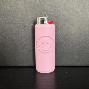 Może przedstawiać: R&oacute;żowa zapalniczka z motywem uśmiechniętej buzi. Zapalniczka ma srebrną metalową g&oacute;rę i czerwony przycisk. Uśmiechnięta buzia jest wytłoczona na r&oacute;żowym korpusie zapalniczki.