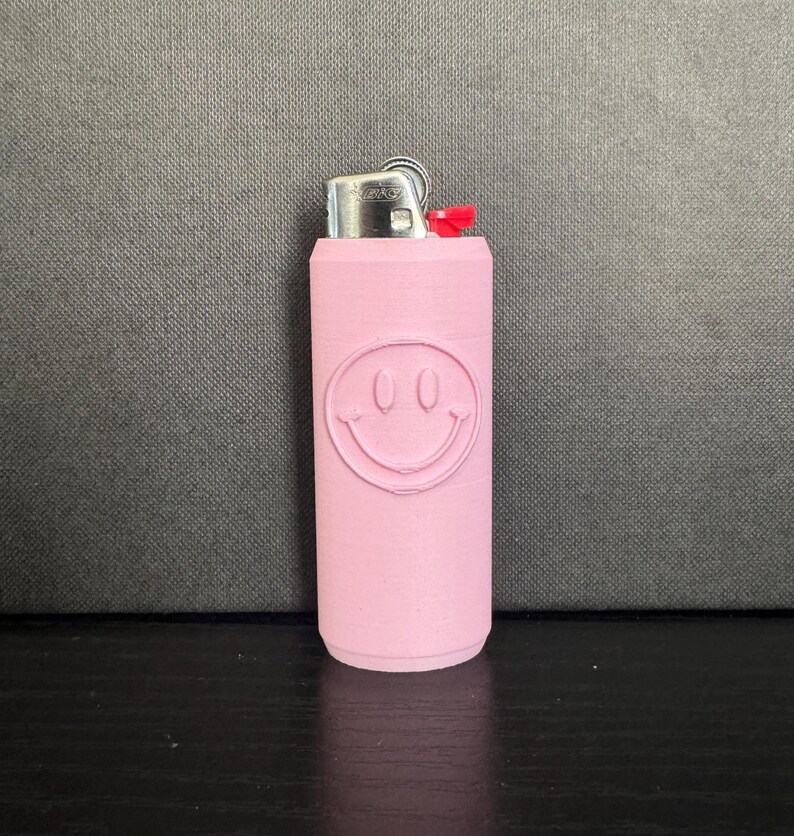 Może przedstawiać: R&oacute;żowa zapalniczka z motywem uśmiechniętej buzi. Zapalniczka ma srebrną metalową g&oacute;rę i czerwony przycisk. Uśmiechnięta buzia jest wytłoczona na r&oacute;żowym korpusie zapalniczki.