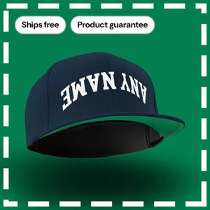 May include: A navy blue baseball cap with a green brim. The cap has white text that says "ANY NAME" on the front.