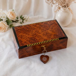 Peut inclure: Une boîte rectangulaire en bois présentant un motif de grain de bois ronceux riche et tourbillonnant sur le couvercle et les côtés. La boîte est ornée d'une bordure rayée noire et jaune et d'un loquet doré avec un porte-clés en bois en forme de cœur.