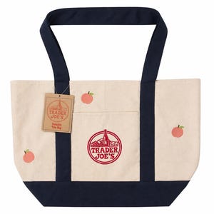 May include: Cream-colored canvas tote bag with navy blue accents. Features the "Trader Joe's" logo in red, peach embroidery, and a hang tag. The bag has a rectangular shape with a navy blue bottom and handles.
