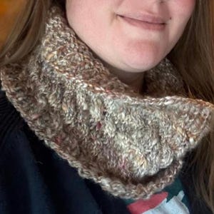 May include: A hand-knitted cowl in shades of brown, beige, and flecks of red and pink. The textured knitwear is worn around the neck, offering warmth and style. The cowl is a cozy accessory for cold weather.
