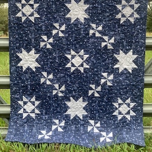 May include: A navy blue and white quilt with a star pattern. The quilt features large white stars and smaller starburst designs on a dark blue background. The quilt is displayed outdoors, draped over a metal fence.