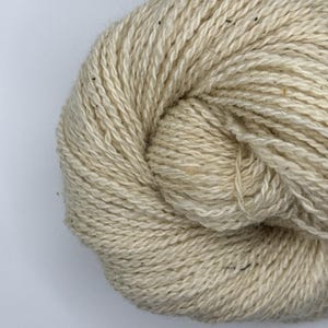 Könnte beinhalten: Nahaufnahme eines Knäuels naturbelassener Wolle. Die Wolle ist hellbeige mit dunkleren Fasern. Das Garn ist eng gewickelt und zeigt die Textur der Fasern. Ideal für Strick- und Häkelprojekte.