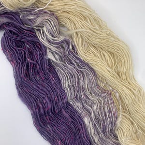 Könnte beinhalten: Nahaufnahme von drei Garnsträngen. Das Garn ist in den Farbtönen Lila, Grau und Creme gehalten. Das lila Garn hat rosa Flecken. Das Garn ist locker gesponnen und hat ein strukturiertes Aussehen. Ideal für Strick- und Häkelprojekte.