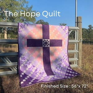 May include: The Hope Quilt, a handmade quilt with a cross design. The quilt features a purple cross on a peach and lavender background, with floral and geometric patterns. Finished size is 142cm x 183cm.