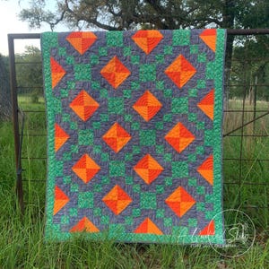 May include: A handmade quilt with a geometric pattern. The quilt features orange diamond shapes on a gray background, interspersed with green square patterns. The border is green, and the quilt is hanging on a metal fence.