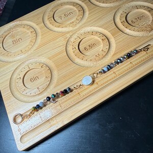 Puede incluir: Una pulsera de cuentas con un cierre dorado y un dije circular blanco descansa sobre una herramienta de medición de pulseras de madera. La pulsera presenta una variedad de cuentas coloridas y un pequeño dije de media luna. La herramienta de medición tiene recortes circulares con medidas en pulgadas.