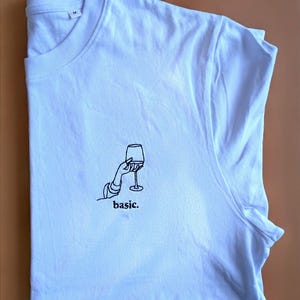 Peut inclure: T-shirt blanc avec un dessin au trait noir d'une main tenant un verre à vin. Le mot "basic" est imprimé en dessous. Le t-shirt est plié sur une surface marron clair.