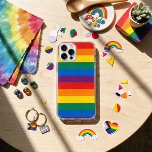 Puede incluir: Una funda de teléfono a rayas arcoíris es el punto focal de esta imagen, rodeada de accesorios con temática Pride. También se ven un paño teñido anudado, pegatinas de arcoíris y un plato pequeño con "Pride 2024". Los pendientes de aro dorados con dijes de la bandera del Orgullo completan el tema.