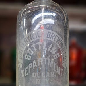 May include: A clear glass soda siphon with a metal top mechanism. The bottle features etched text reading DOTTERWEICH BREWING CO. BOTTLING DEPARTMENT OLEAN, N.Y. The metal nozzle is silver-toned and sits atop the cylindrical glass vessel.