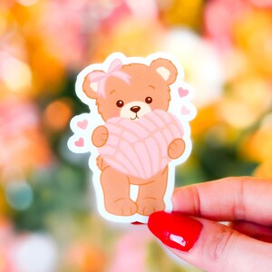 May include: A sticker featuring a cartoon bear holding a pink concha pastry. The bear is light brown with a pink bow and small hearts. The background is a blurred mix of pink, yellow, and orange.