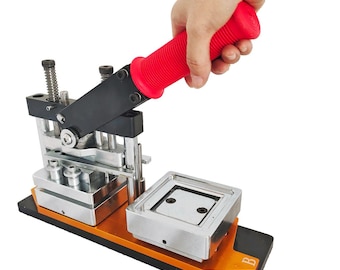 May include: A red-handled badge making machine with a metal base and a silver-colored die. The machine has a lever that is being pressed down by a hand.