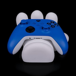 May include: A blue and white Xbox controller resting on a white stand, set against a black background. The controller has a sleek design with black buttons and a central Xbox logo. The stand has a unique, curved shape.