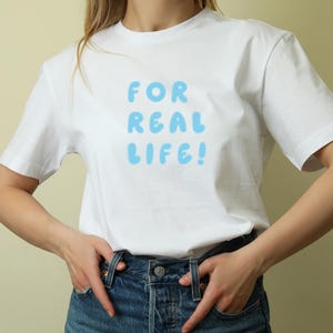 Puede incluir: Camiseta blanca con el texto "FOR REAL LIFE!" en letras de burbujas azul claro. La camiseta se combina con vaqueros azules. El estilo general es informal y relajado.