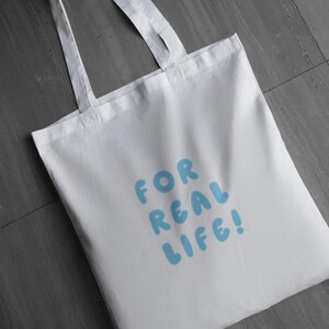 Puede incluir: Bolso tote blanco con el texto "FOR REAL LIFE!" en letras de burbujas azul claro. El bolso tiene dos asas blancas y est&aacute; hecho de una tela lisa y sin adornos. El bolso est&aacute; sobre una superficie gris.