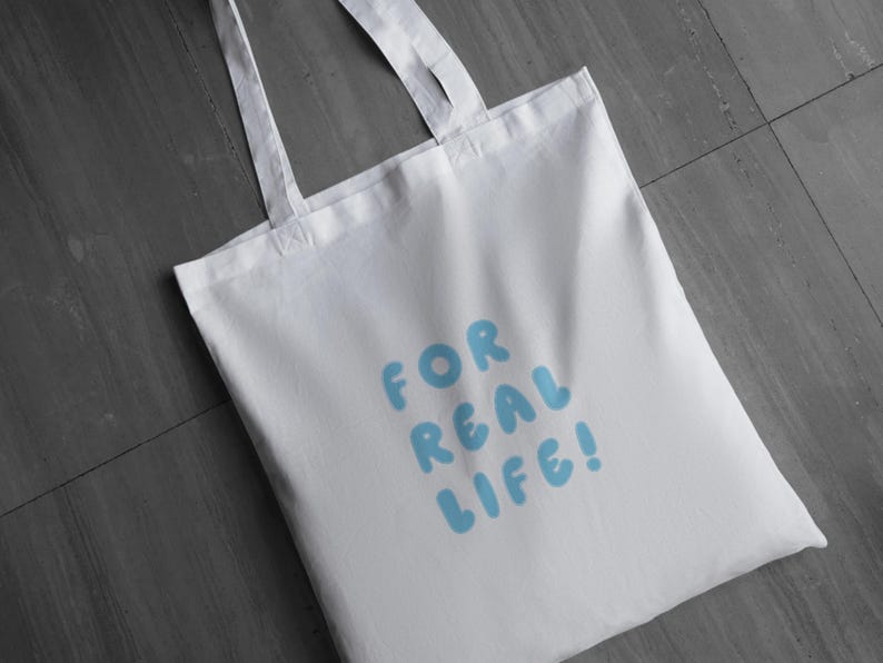 Puede incluir: Bolso tote blanco con el texto "FOR REAL LIFE!" en letras de burbujas azul claro. El bolso tiene dos asas blancas y est&aacute; hecho de una tela lisa y sin adornos. El bolso est&aacute; sobre una superficie gris.