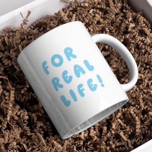 Puede incluir: Una taza de cer&aacute;mica blanca con el texto "FOR REAL LIFE!" en letras de burbujas azul claro. La taza est&aacute; dentro de una caja blanca llena de papel arrugado marr&oacute;n.