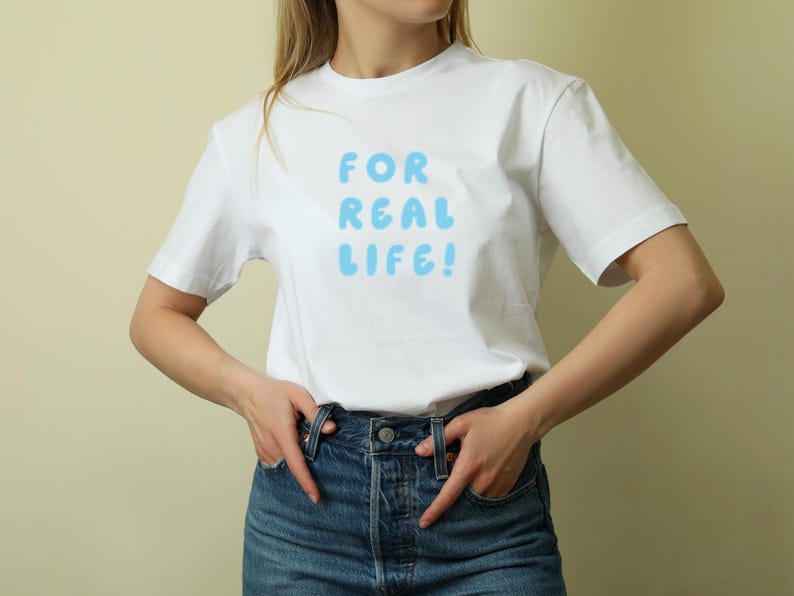 Puede incluir: Camiseta blanca con el texto "FOR REAL LIFE!" en letras de burbujas azul claro. La camiseta se combina con vaqueros azules. El estilo general es informal y relajado.