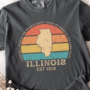 May include: Dark gray t-shirt featuring a retro design with the state of Illinois in a tan color, set against a striped background of orange, yellow, and blue. The text includes "ILLINOIS EST 1818" and state slogans.