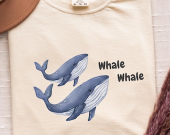 May include: Cream-colored t-shirt featuring two blue watercolor whales and the text "Whale Whale." The shirt has a classic crew neck and a tag that reads "XL-TG-EG."