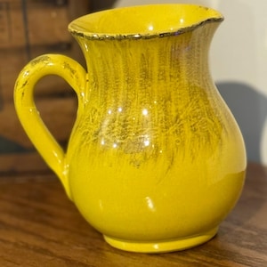 May include: A yellow ceramic pitcher with a curved handle. The pitcher has a textured surface with darker yellow and gold accents. The rim is outlined with a gold trim. The pitcher is sitting on a wooden surface.