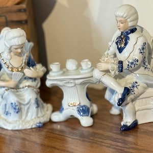 May include: A porcelain figurine set depicting a man and woman in 18th-century attire, seated near a small table with teacups. The figures are predominantly white with blue floral accents and gold trim. The table is white with blue accents.