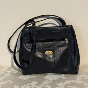 May include: Black leather shoulder bag with a top handle and a long strap. The bag has a front zipper pocket and a gold-colored oval emblem. The bag is sitting on a patterned surface.