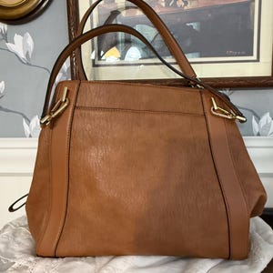 May include: A brown handbag with two handles and gold-colored hardware. The bag has a textured appearance and a rectangular shape with a slightly trapezoidal profile. The handles are attached with gold-colored metal pieces.