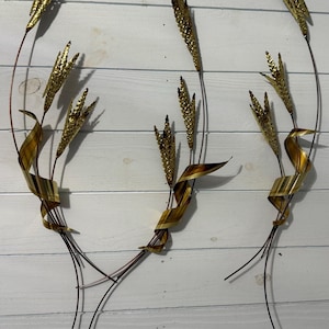 Puede incluir: Arte mural de metal que presenta tres espigas de trigo estilizadas con cabezas doradas y tallos marrones. Las espigas de trigo tienen acentos en forma de cinta marrón y dorada. La obra de arte está montada sobre un fondo de madera blanca.