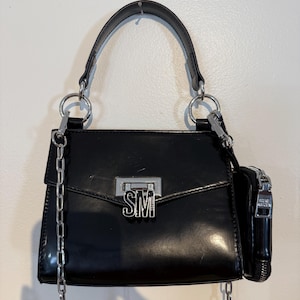 May include: Black handbag with a silver chain strap and a small attached pouch. The bag has a top handle and a silver clasp with the letters "SM". The bag is hanging against a white wall.