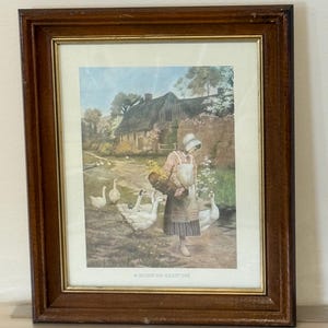 May include: Framed print of a rural scene with a woman in a bonnet carrying a basket and a goose, surrounded by geese. The artwork is set in a wooden frame with a gold inner border. The print has a vintage aesthetic.