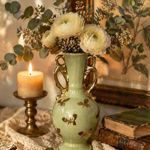 May include: A light green vase with gold handles and butterfly designs holds a bouquet of white flowers and greenery. A lit candle in a gold candlestick and two antique books are also visible, creating a vintage aesthetic.