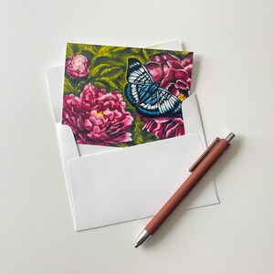 May include: A greeting card with a painting of a blue and white butterfly on a pink flower, inside a white envelope. A brown pen lies nearby. The card's artwork depicts pink peonies and green leaves.
