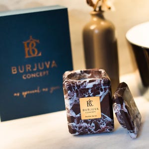 Può includere: Una candela in marmo marrone e bianco con un'etichetta dorata che recita "Burjuva Concept Marble Series". La candela è in un contenitore quadrato con un coperchio abbinato.