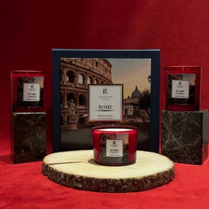 Puede incluir: Exhibición de velas de vidrio rojo "ROME" con una imagen del Coliseo. Las velas están sobre bloques de mármol y una rodaja de madera, con una caja con el mismo tema en el fondo. Las velas son de "BURJUVA CONCEPT".
